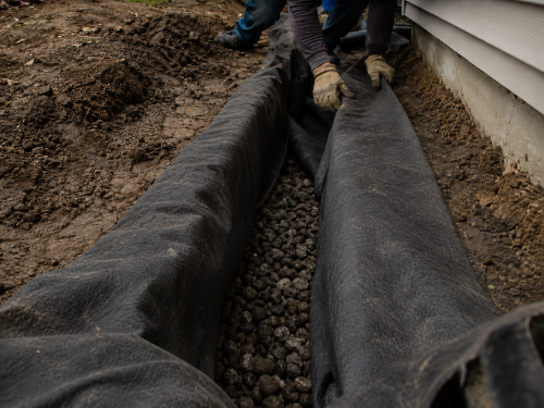 Worker wrapping non-woven geotextile drainage fabric over a gravel foundation French drain
