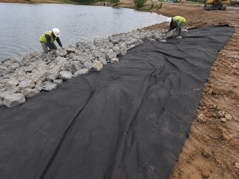 Workers installing a large black landscape fabric liner along the edge of a rocky lake or pond on a construction site.